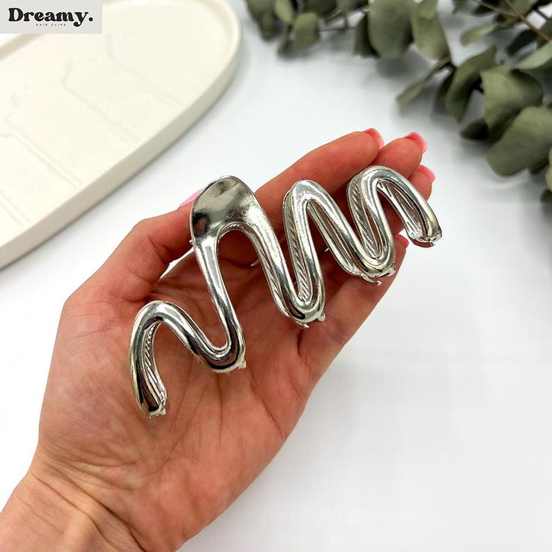 May include: A silver-colored hair clip in a wavy, abstract design. The clip is held in a hand, showcasing its polished, metallic finish. The clip's design is reminiscent of a stylized snake or wave, adding a modern touch to hair accessories.