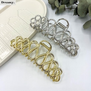 May include: Two metal hair claw clips. One is gold-toned, the other is silver-toned. Both have an open, abstract design with a spring hinge. The clips are on a white surface.