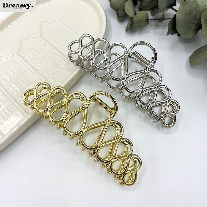 May include: Two metal hair claw clips. One is gold-toned, the other is silver-toned. Both have an open, abstract design with a spring hinge. The clips are on a white surface.