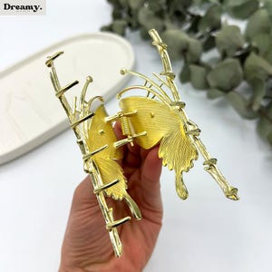 May include: Gold-colored butterfly hair claw clip. The clip features a detailed butterfly wing design with a textured finish. The clip is made of metal and has a spring mechanism for securing hair. The clip is a hair accessory.
