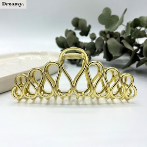 May include: A gold-coloured hair claw clip with an open, looped design. The clip has a spring mechanism and is designed to hold hair in place. The word "Dreamy" is in the top left corner.