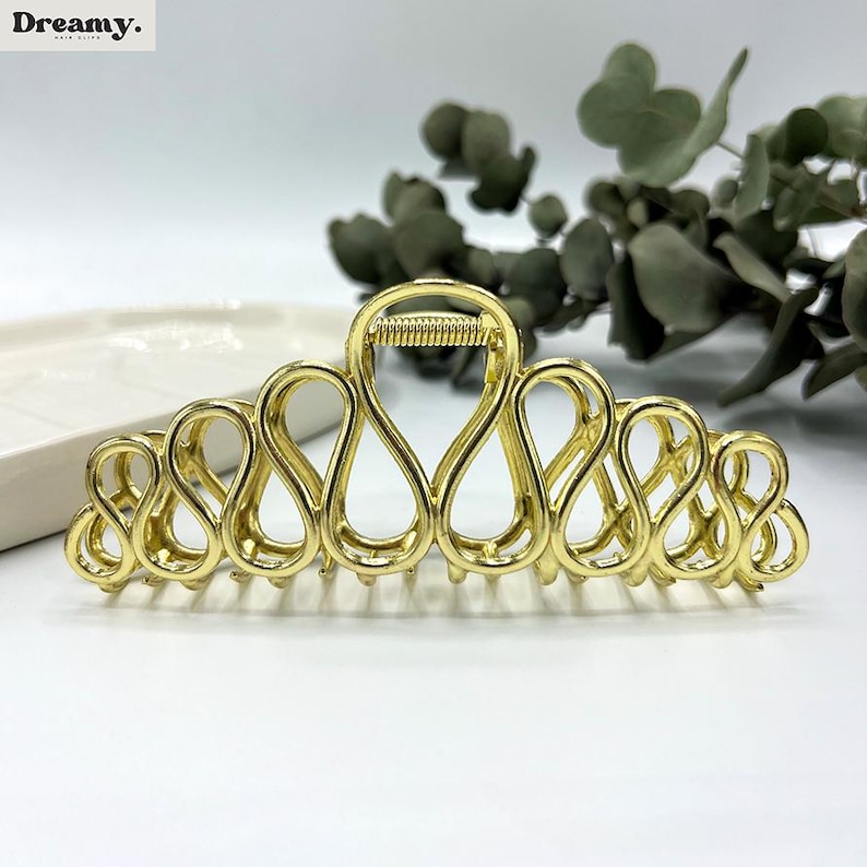 May include: A gold-coloured hair claw clip with an open, looped design. The clip has a spring mechanism and is designed to hold hair in place. The word "Dreamy" is in the top left corner.