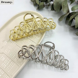 May include: Two metal hair claw clips. One is gold-coloured, the other is silver-coloured. Both have an open, looped design. The clips are on a white tray with a textured surface, with greenery in the background. The brand name "Dreamy" is in the top left corner.
