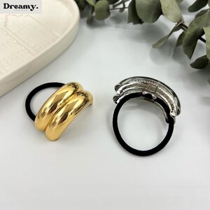 May include: Two hair ties, one gold and one silver, with a black elastic band. The gold hair tie is a double band, while the silver hair tie is a single band.