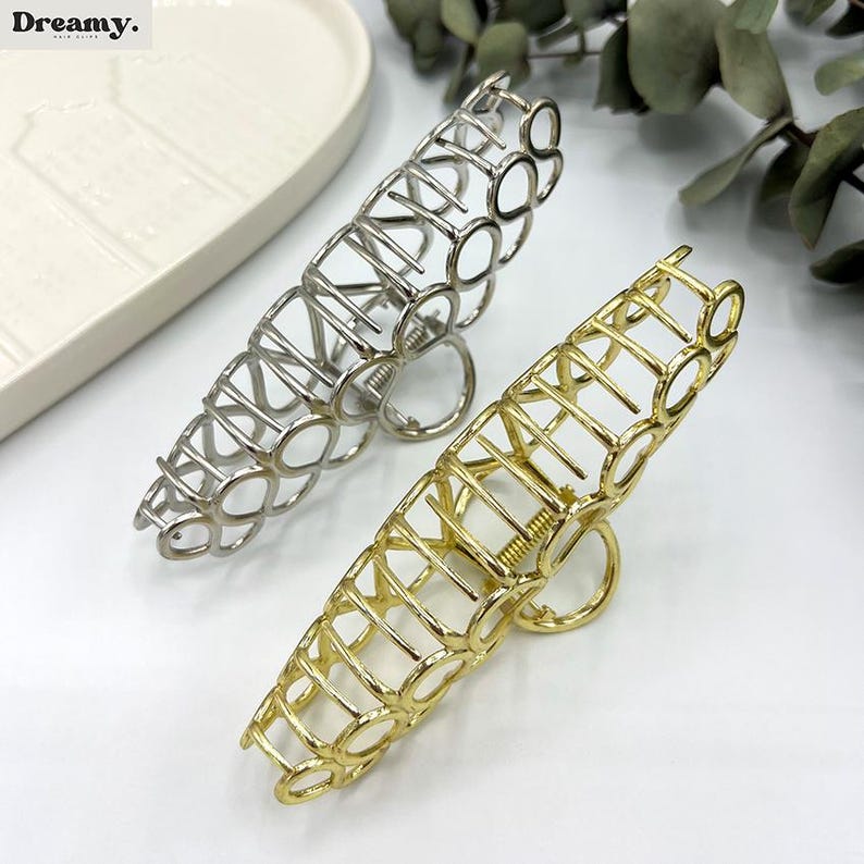 May include: Two metal hair claw clips, one silver and one gold, are shown. The clips feature an open, geometric design with a circular pattern. The word "Dreamy" is visible in the top left corner.
