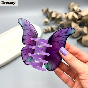 May include: A purple butterfly-shaped hair claw clip. The clip features detailed wing designs in shades of purple and teal. The clip's clasp is a matching lavender colour. The word "Dreamy" is printed in white in the upper left corner.