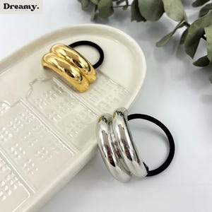 May include: Two hair scrunchies, one gold and one silver, with a double-ring design. The scrunchies are on a white ceramic plate with a raised pattern.
