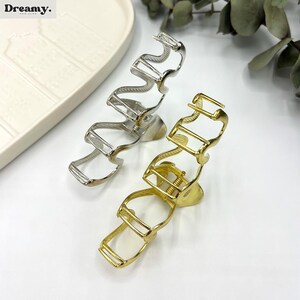 May include: Two metal hair claw clips. One is silver, the other is gold. The silver clip has a wavy design, while the gold clip has a rectangular design. The clips are on a white surface. The brand name "Dreamy" is in the top left corner.