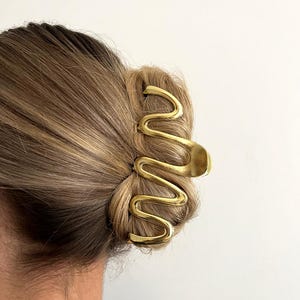 May include: A gold-colored hair clip in a wavy design, holding up a bun. The clip is made of metal and has a smooth, shiny finish. The word "Dreamy" is printed in black in the upper right corner.