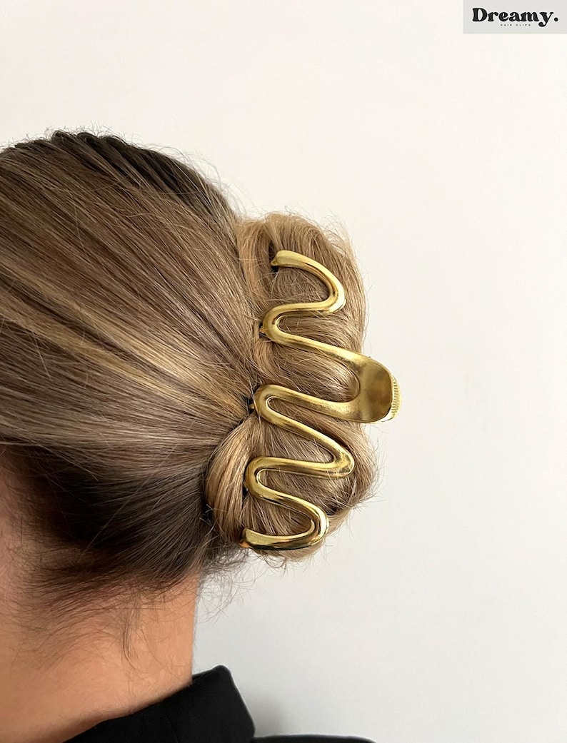 May include: A gold-colored hair clip in a wavy design, holding up a bun. The clip is made of metal and has a smooth, shiny finish. The word "Dreamy" is printed in black in the upper right corner.