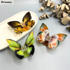 May include: Three butterfly hair clips in yellow, green, and orange. The clips feature detailed wing patterns and clear clasps. One clip sits on a white, oval dish. The Dreamy logo is in the top left corner.