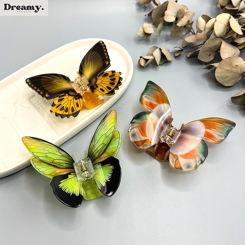 May include: Three butterfly hair clips in yellow, green, and orange. The clips feature detailed wing patterns and clear clasps. One clip sits on a white, oval dish. The Dreamy logo is in the top left corner.