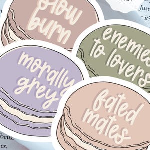 May include: A collection of four round stickers with text. The stickers are in pastel colors: mauve, sage green, lavender, and peach. The text on the stickers reads: "slow burn", "enemies to lovers", "morally grey", and "fated mates".