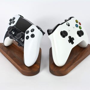 May include: Two white and black game controllers on individual wooden stands. One controller has a black center and black buttons. The other controller has colored buttons. The stands are a dark brown color.