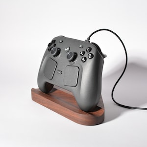 May include: A dark gray video game controller with two analog sticks, a directional pad, four lettered action buttons, and two touchpads, resting on a curved dark wood stand. A black cable extends from the top of the controller to the right.