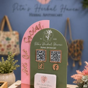 Könnte beinhalten: Ein zweilagiges Acrylschild mit einer rosafarbenen Bogenplatte mit dem roségoldenen Text Let's Get Social und einer salbeigrünen Bogenplatte mit zwei QR-Codes, TikTok- und Facebook-Symbolen sowie dem Text Pita's Herbal Haven Herbal Apothecary.