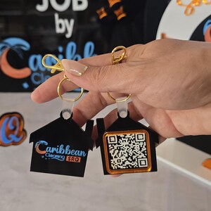 May include: Two black house-shaped keychains with gold-colored hardware. One keychain has the text "Caribbean Cleaning SRQ" and a logo. The other has a QR code. The background includes text that says "ANOTHER SPARKLING JOB".