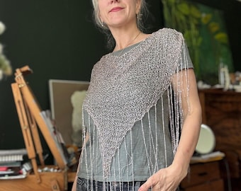Könnte beinhalten: Ein silberner Perlen-Schal mit langem Fransenbesatz wird über einem grauen T-Shirt und einer dunkel gewaschenen Jeans getragen. Der Schal ist dreieckig und scheint handgestrickt zu sein. Die Frau im Bild blickt nach oben und nach links.
