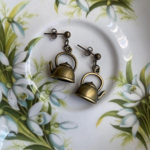 Könnte beinhalten: Ein Paar goldfarbene Teekannen-Ohrringe mit einem baumelnden Anhänger. Die Ohrringe befinden sich auf einem weißen Hintergrund mit einem Blumenmuster.