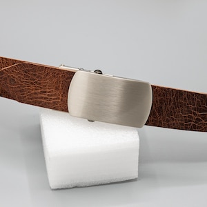 May include: A brown leather belt with a silver-colored buckle. The belt has a textured surface and is displayed against a white background. The buckle is rectangular with rounded edges.