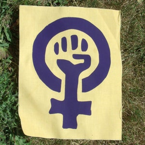 May include: A purple feminist symbol with a raised fist inside the circle on a yellow background.