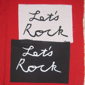 May include: Two fabric patches with the phrase "Let's Rock" written in black and white. The top patch is pink and the bottom patch is black.