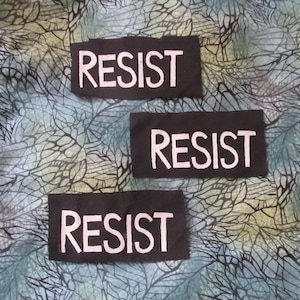 May include: Three black fabric patches with white text that reads "RESIST" on a blue and green patterned background.