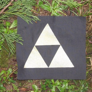 May include: A black fabric patch with a white triangular symbol, resembling the Triforce from the Legend of Zelda video game series. The patch is lying on a bed of green grass and brown leaves.