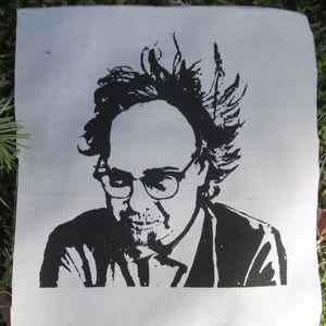 May include: Black and white image of a person with glasses and messy hair wearing a dark suit jacket. The image is printed on a white fabric patch.