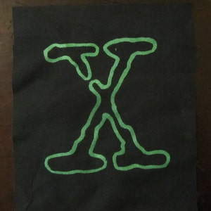 May include: A black fabric patch with a green outline of the letter "X" in the center.