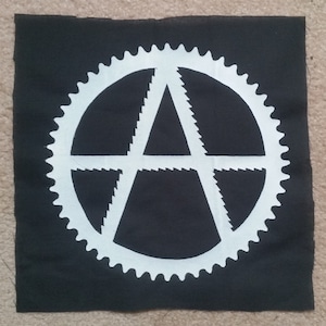 May include: Black fabric patch with a white anarchy symbol inside a white cog.