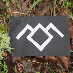 Peut inclure: Un symbole blanc sur un patch de tissu noir. Le symbole est une chaîne de montagnes stylisée avec un diamant au centre.