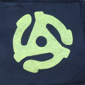 May include: A stack of black fabric squares with a neon green graphic of a 45 RPM record adapter printed on each. The edges of the fabric are frayed, giving a rustic appearance. The background is a weathered wooden surface.