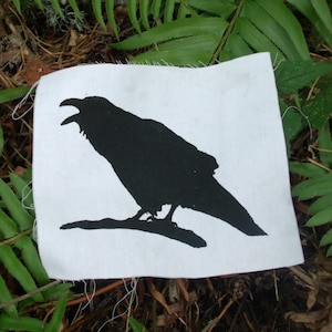 May include: A black silhouette of a raven on a white fabric patch. The raven is perched on a branch with its wings spread out.