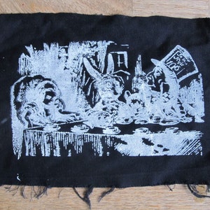 May include: A black fabric patch with a white image of a tea party scene from Alice in Wonderland. The Mad Hatter is wearing a top hat with the words "Drink Me" on it.
