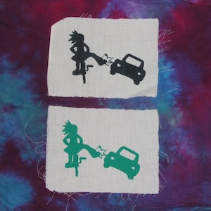 Op de afbeelding: Twee stoffen patches met een zwart-witte afbeelding van een persoon die een auto trapt op de ene patch en een groen-witte afbeelding van een persoon die een auto trapt op de andere patch.