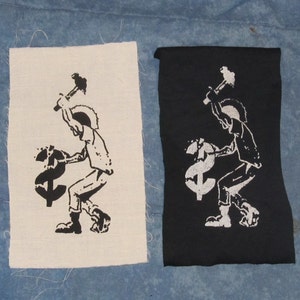 May include: Two fabric patches with a black and white image of a person smashing a dollar sign with a hammer. The patches are on a blue fabric background.