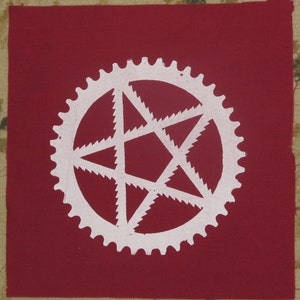 May include: A white five-pointed star with serrated edges is centred inside a white gear-shaped circle. The design is on a red fabric background.
