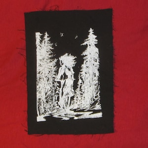 May include: Black and white embroidered patch with a stylized image of a person standing in a forest. The person is facing to the right and has long hair. The trees are tall and slender. The patch has a frayed edge.