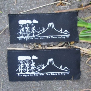 Puede incluir: Dos parches rectangulares negros con ilustraciones blancas de un paisaje con montañas, nubes y mazorcas de maíz. El texto "Sin Agua no hay Vida Sin Tierra no hay Paz" está impreso en cada parche.
