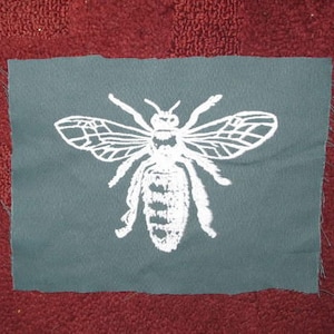 May include: A white embroidered bee design on a dark grey fabric patch.