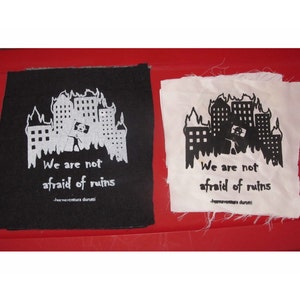 May include: Two black and white fabric patches with a graphic of a burning city and the text "We are not afraid of ruins -aventura durutti".