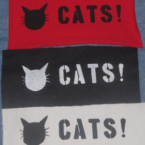 Puede incluir: Tres paneles de tela con la palabra "CATS!" estampada en negro, blanco y gris sobre un fondo rojo, negro y blanco. Cada panel presenta una silueta negra, blanca o gris de una cabeza de gato.