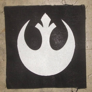 Puede incluir: Un parche de tela negro con un símbolo blanco de la Alianza Rebelde. El símbolo es un pájaro estilizado con las alas extendidas, que se asemeja a una luna creciente.