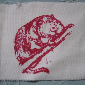 Op de afbeelding: Een rode geprinte afbeelding van een klein dier, mogelijk een eekhoorn of een chipmunk, op een witte stoffen achtergrond. Het dier wordt afgebeeld terwijl het een tak beklimt.