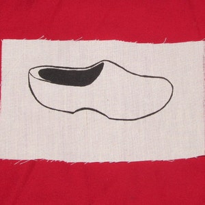 May include: A white wooden shoe with a black outline, on a white fabric background with a red border.
