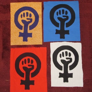 May include: Four fabric patches with a feminist symbol, a circle with a raised fist inside, on a red, white, blue, and yellow background.