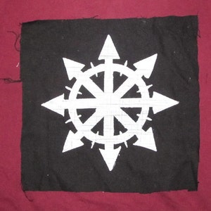 May include: A black fabric patch with a white eight-pointed star with arrows pointing outward. The star has a circle in the center with eight spokes.