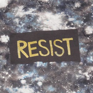 May include: A black fabric patch with the word "RESIST" written in gold glitter letters against a galaxy-print background.