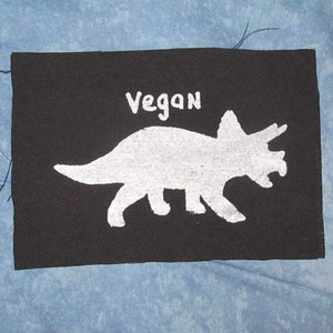 May include: Black fabric patch with a white silhouette of a dinosaur and the word "Vegan" written in white.